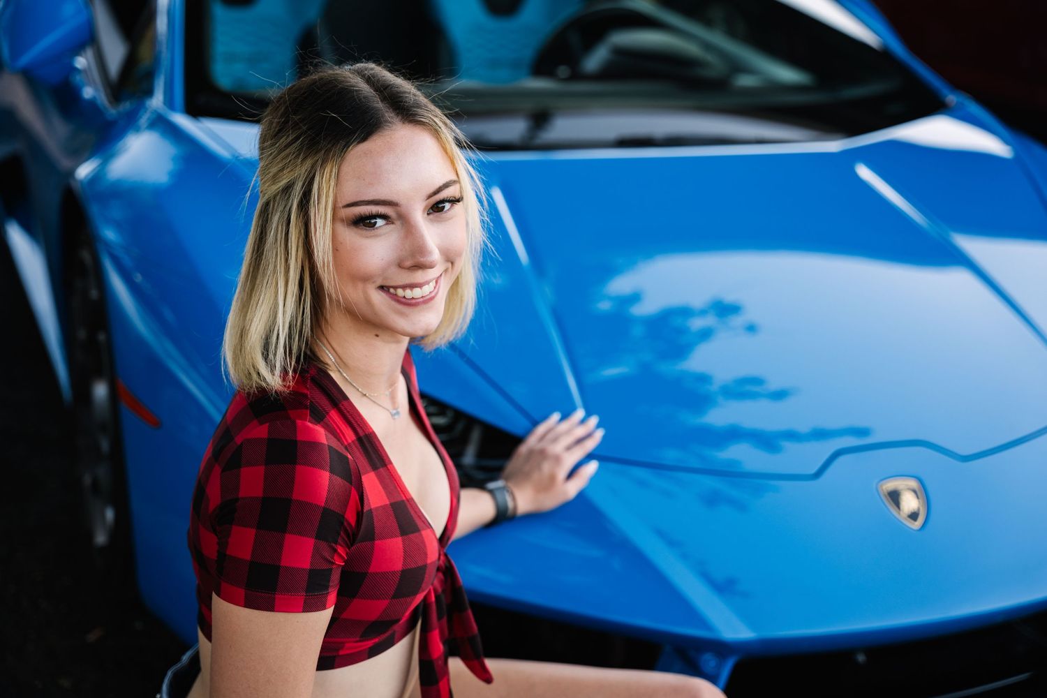 Person in plaid crop top stands beside bright blue luxury sports car.