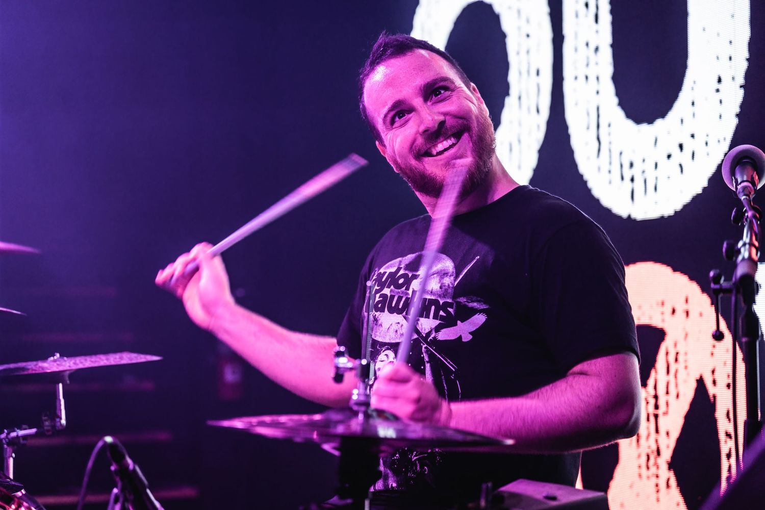 Drummer performing on stage with purple lighting and expressive energy.