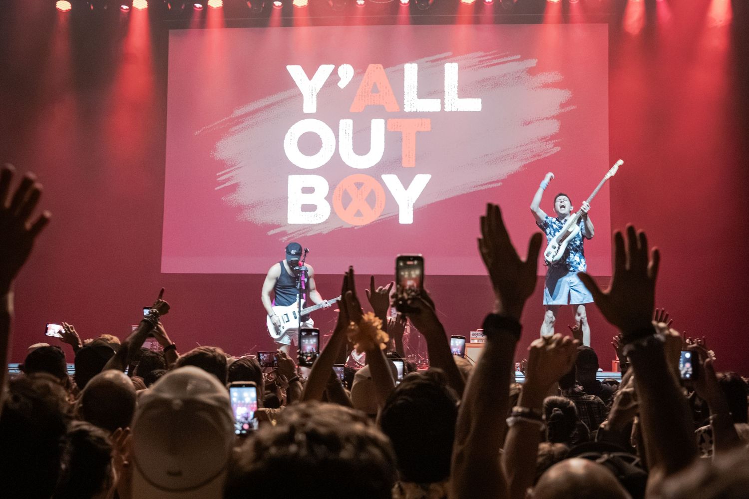 Y'All Out Boy concert with energetic crowd raising hands and phones at a live music event on a red-lit stage.