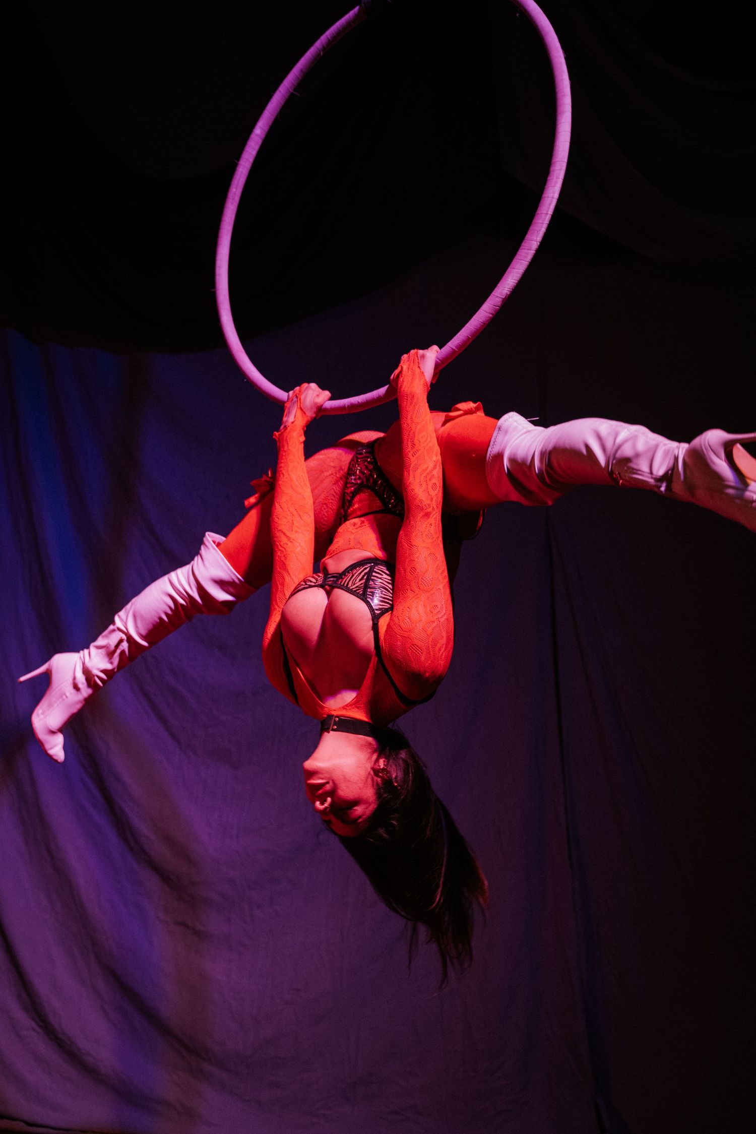 Aerial hoop acrobat performs inverted pose in red lighting during circus performance.