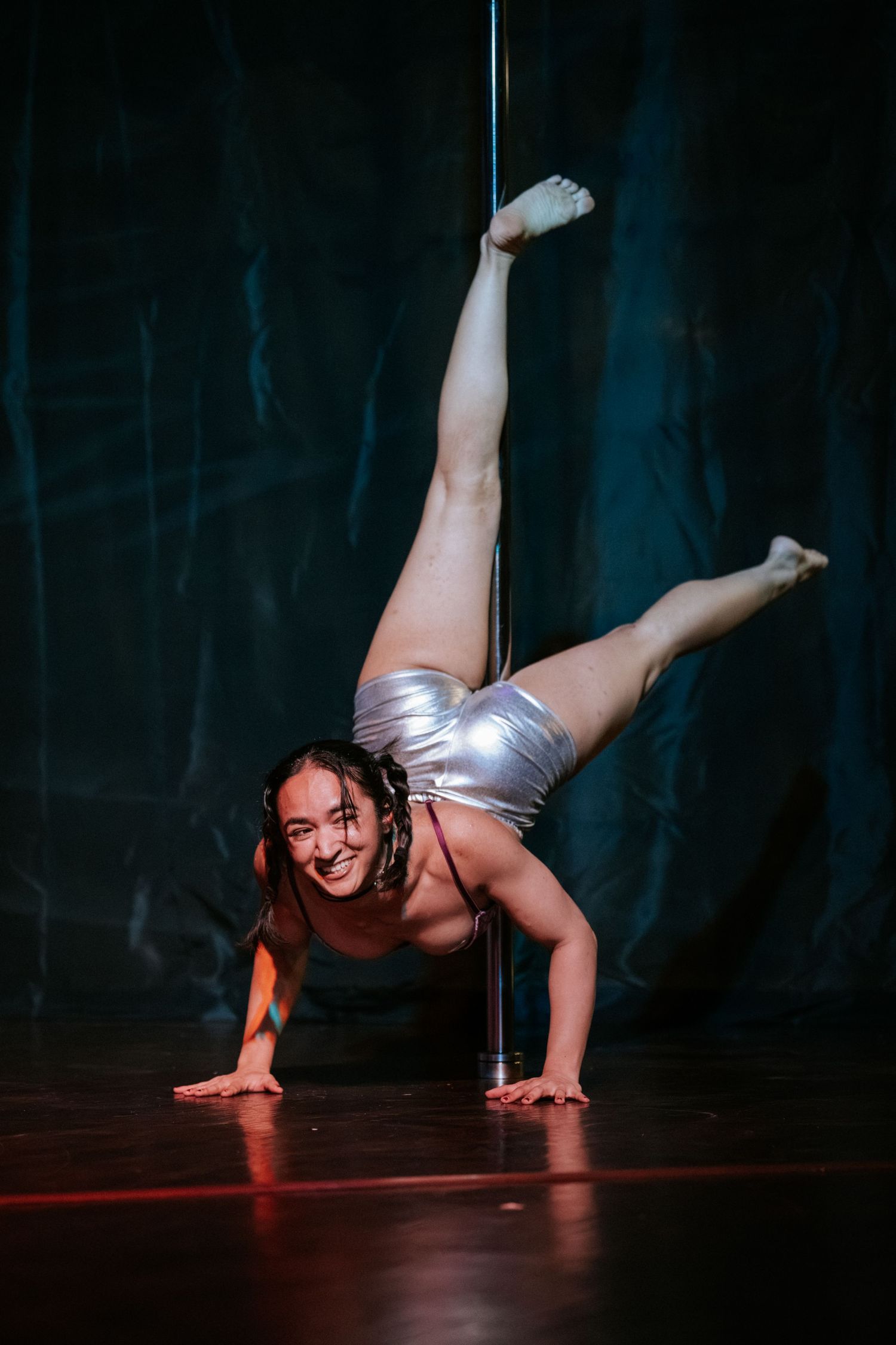 Smiling pole dancer in silver outfit performs handstand split on stage with dark teal backdrop.