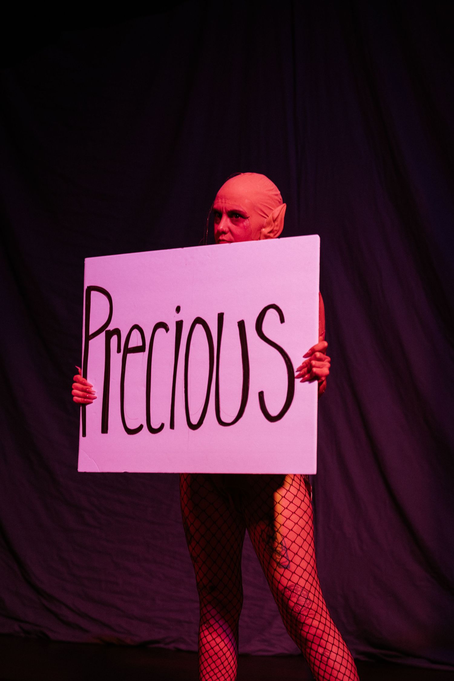 Person in pink bodysuit holding a sign reading Precious on a dark stage under pink lighting.