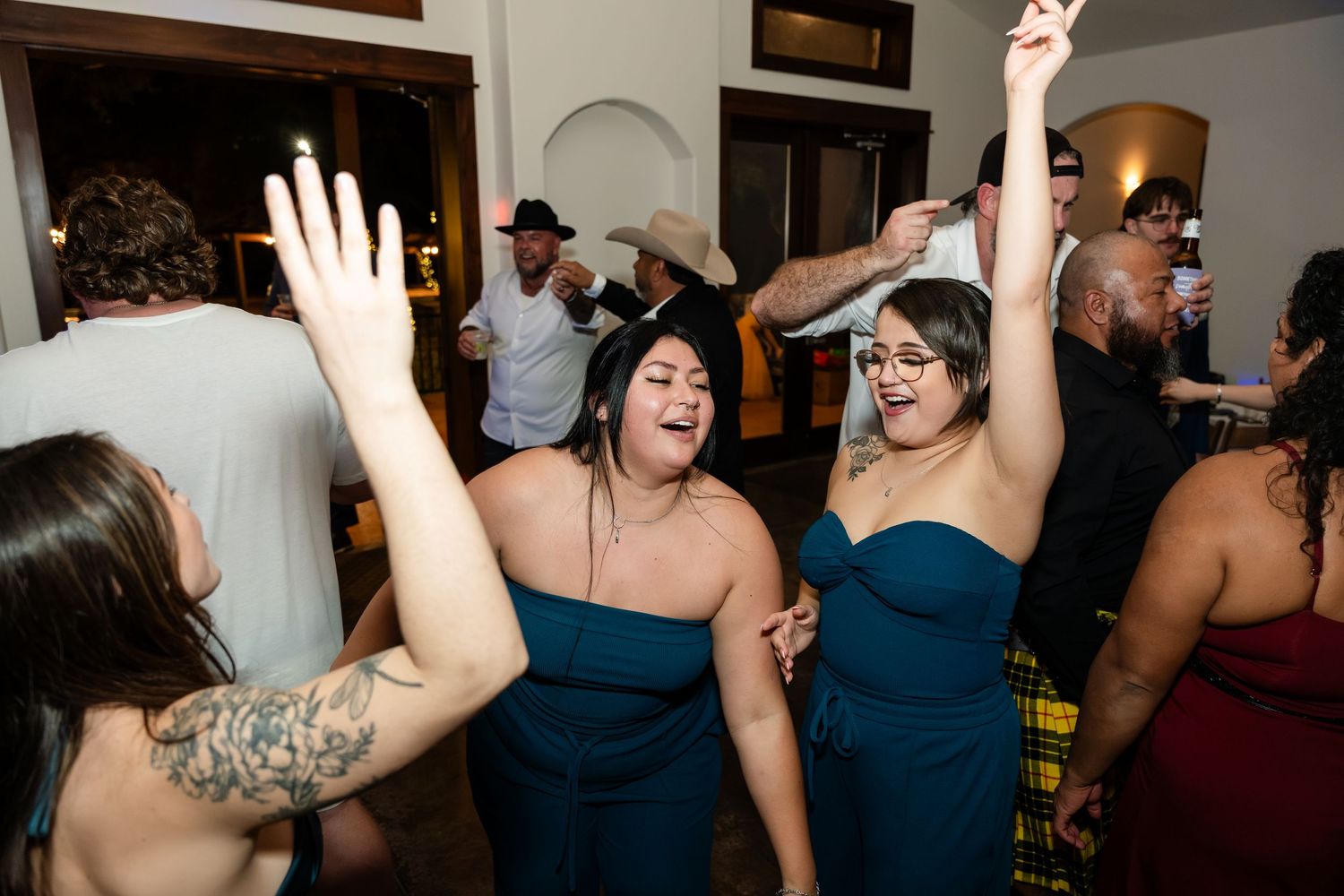 Bridesmaids in teal strapless dresses dance joyfully at a wedding reception with guests celebrating in the background.