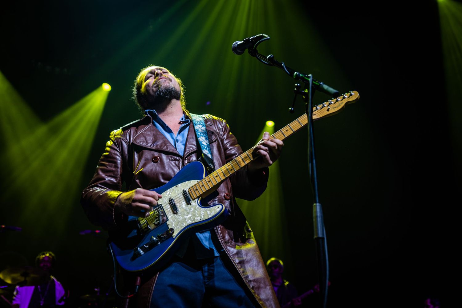 Musicians perform on stage with electric guitars under dramatic green lighting during a concert performance.
