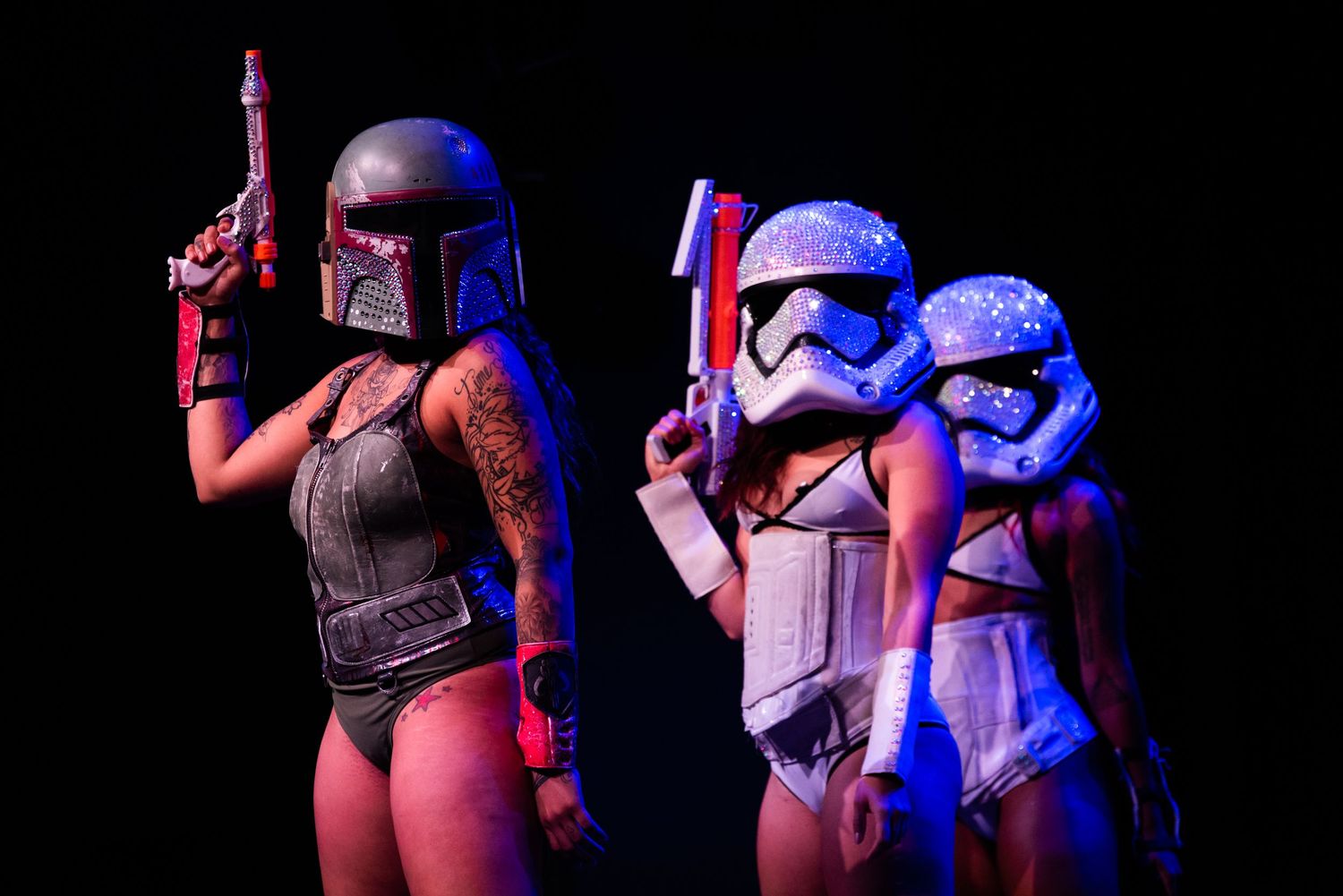 Performers in lingerie and stormtrooper helmets pose dramatically on stage under blue lighting during a burlesque show.