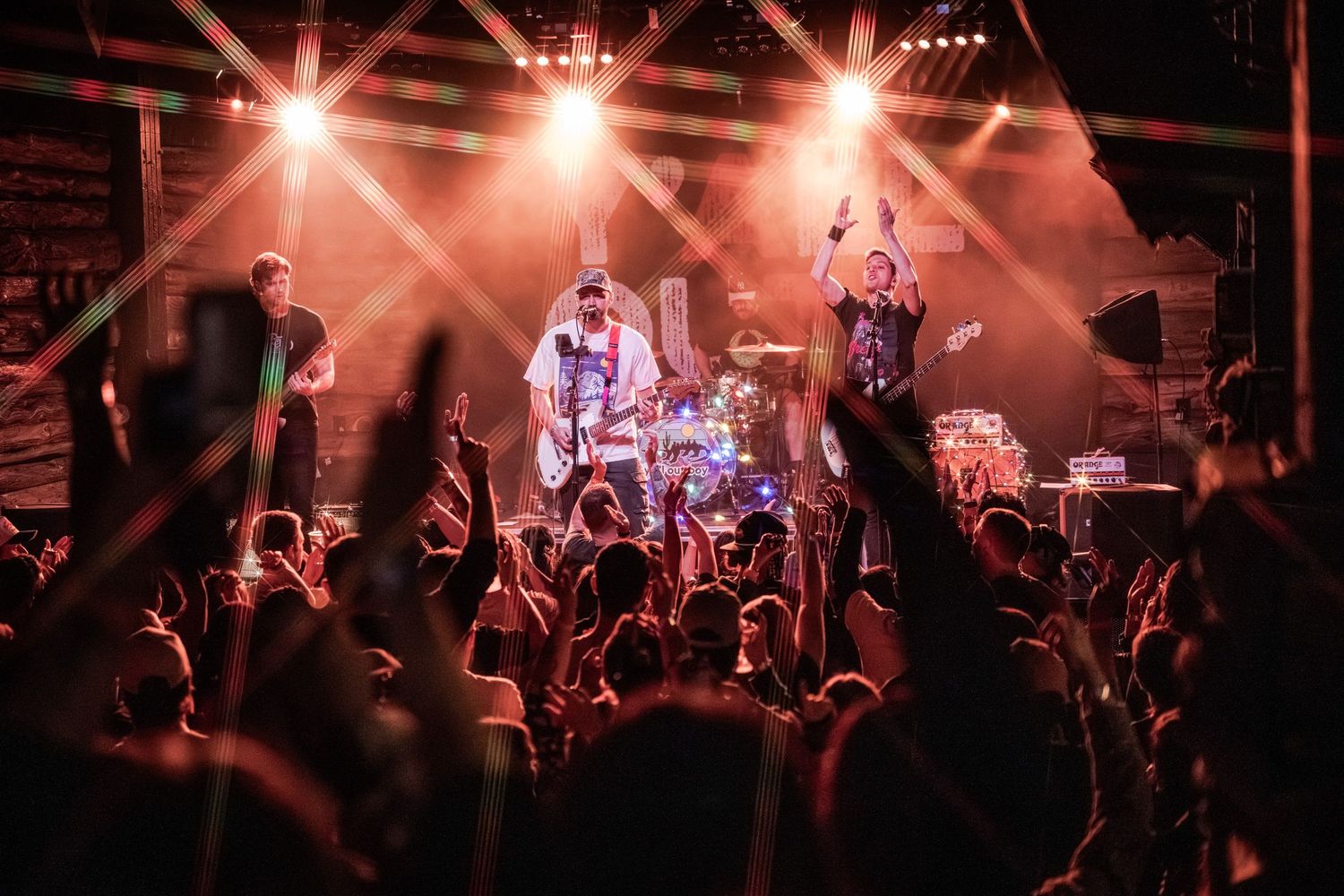 A rock band performs on stage with dramatic red lighting while an energetic crowd cheers in the foreground.