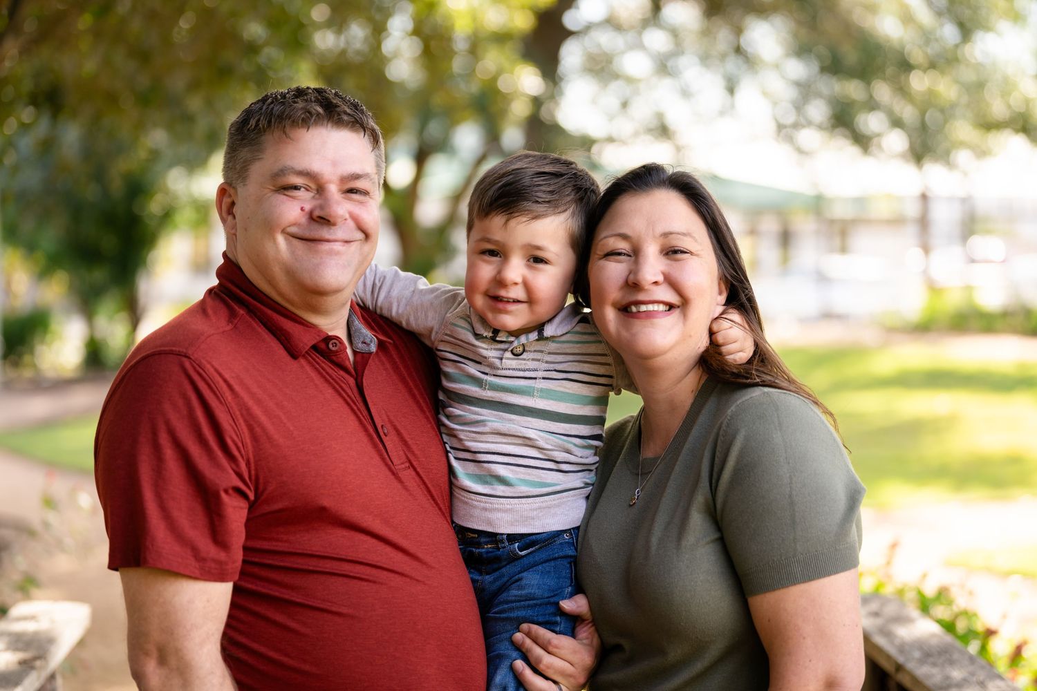 A family of three poses together outdoors on a sunny day in a park setting.