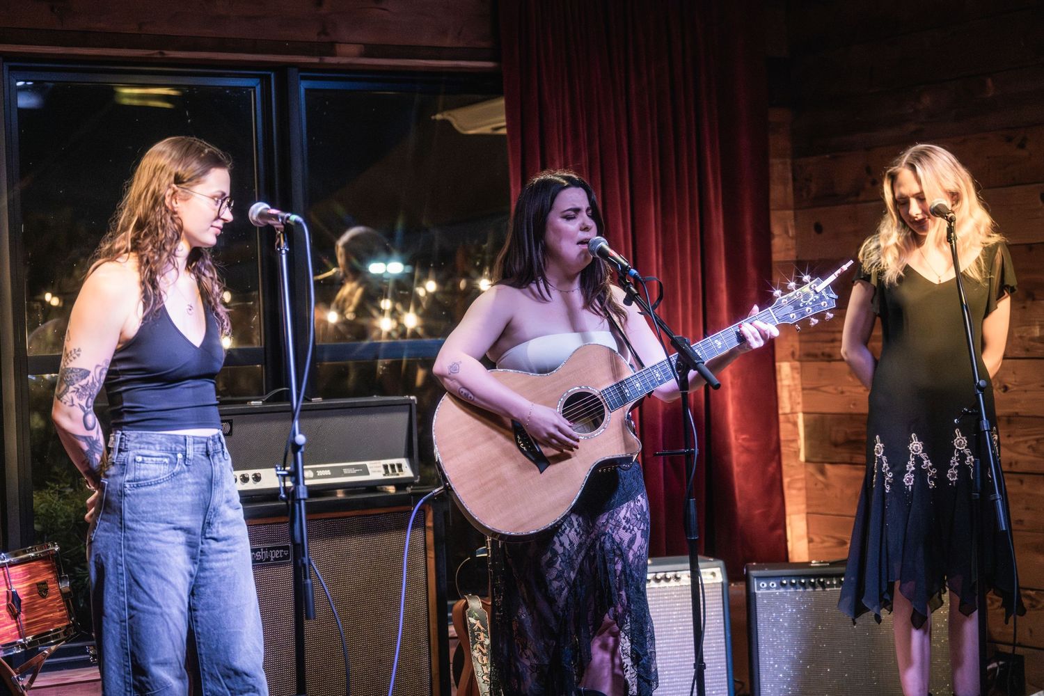 Three performers sing together on stage with an acoustic guitar at an intimate music venue with red curtains.