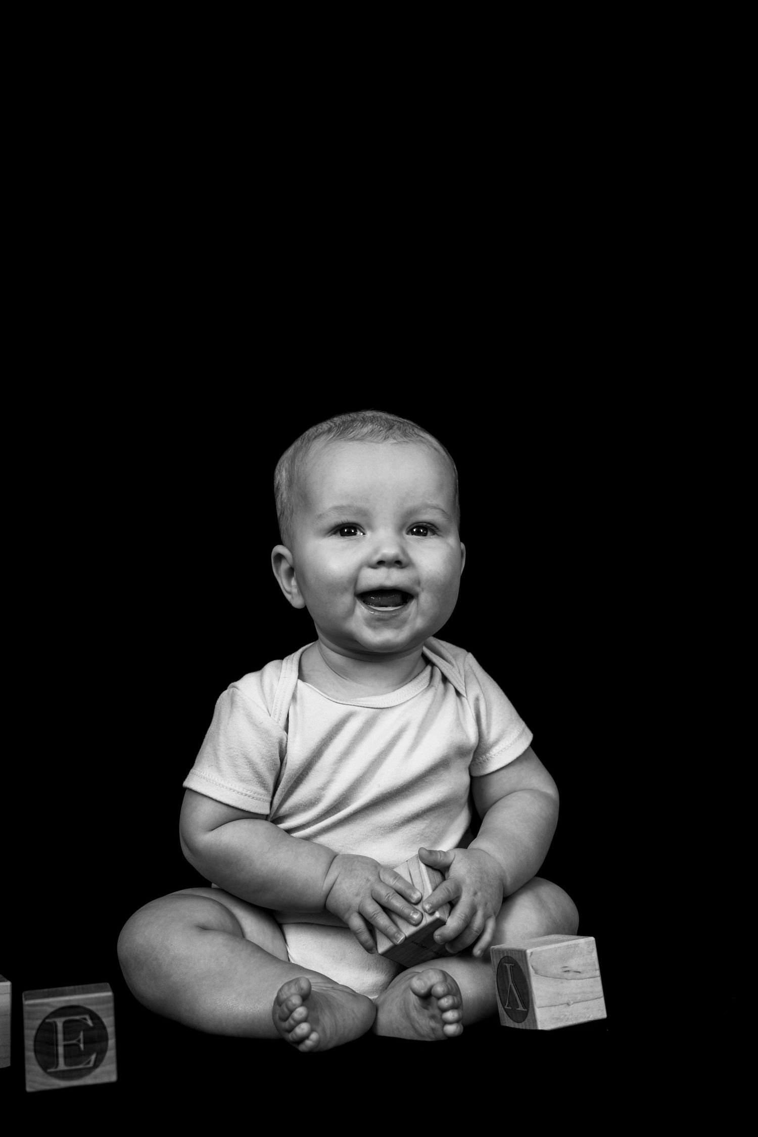 A joyful baby in a white onesie sits against a black background with wooden alphabet blocks.