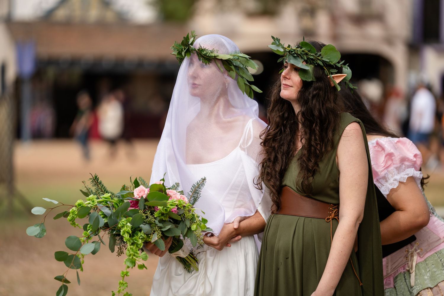A wedding party stands together in an outdoor garden setting wearing olive green and white dresses with floral accents.