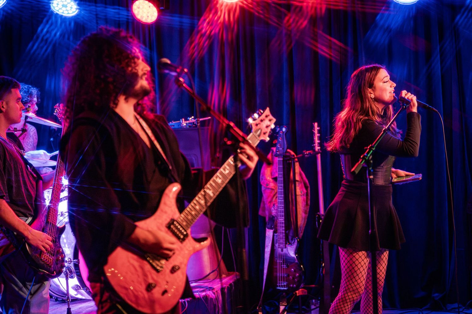 Musicians perform on stage with guitars and microphones under dramatic blue and red lighting during a live concert.