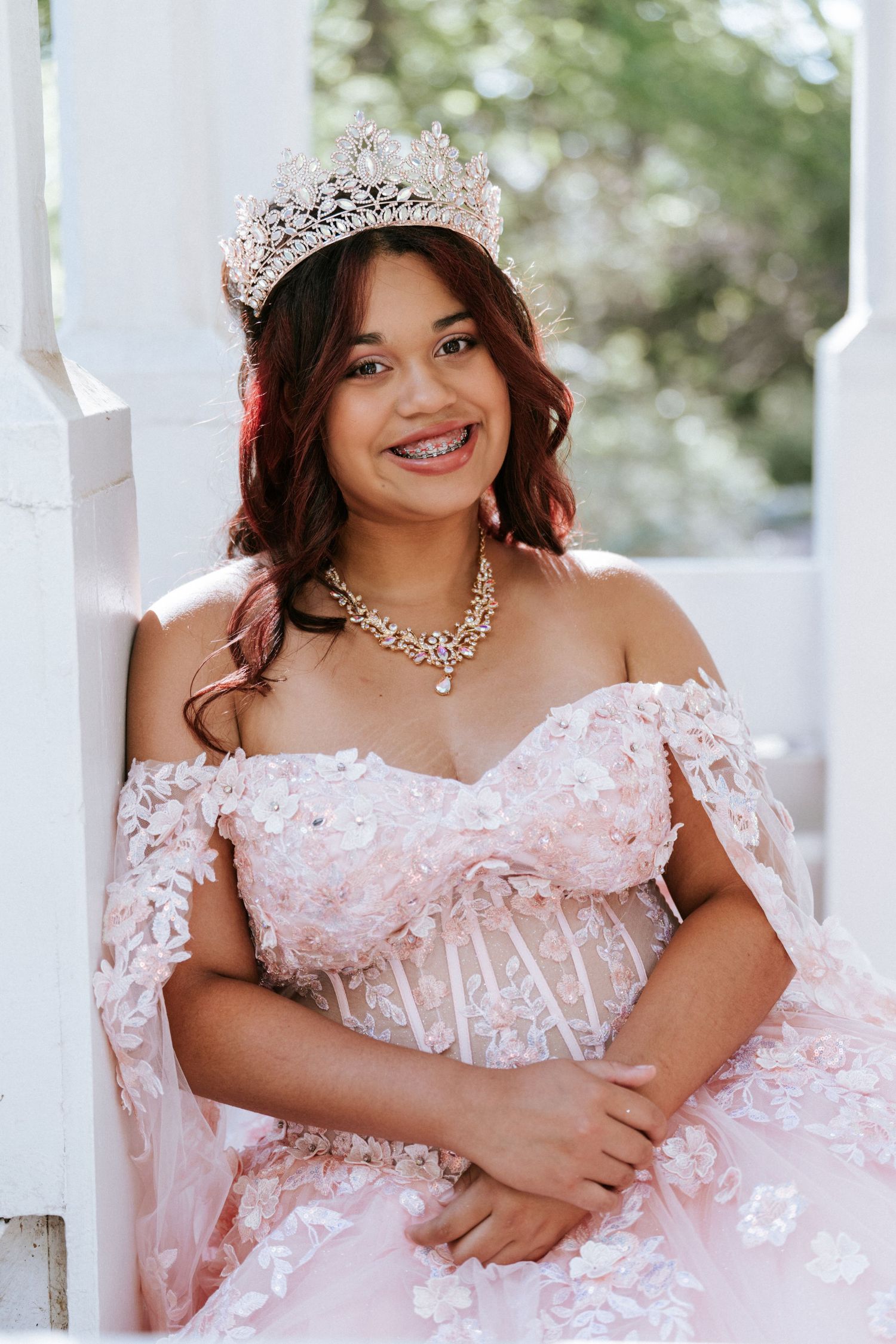 Young woman in pink floral off-shoulder gown, diamond tiara, and statement necklace smiling at quinceañera celebration.