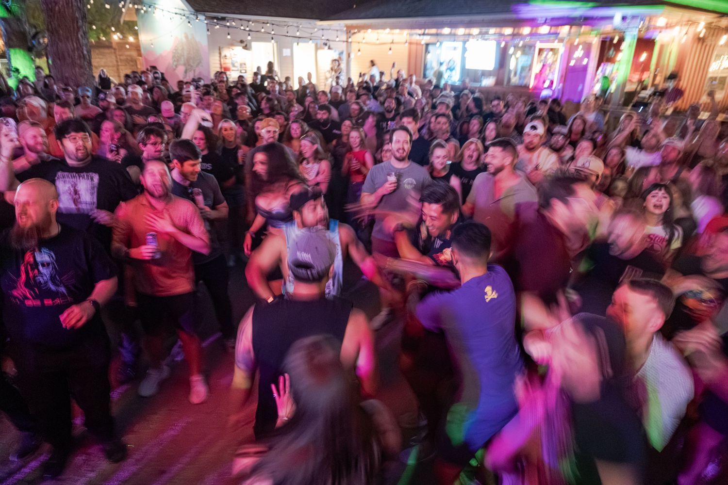 Large crowd of people dancing and celebrating at a vibrant indoor party with colorful purple and green lights.