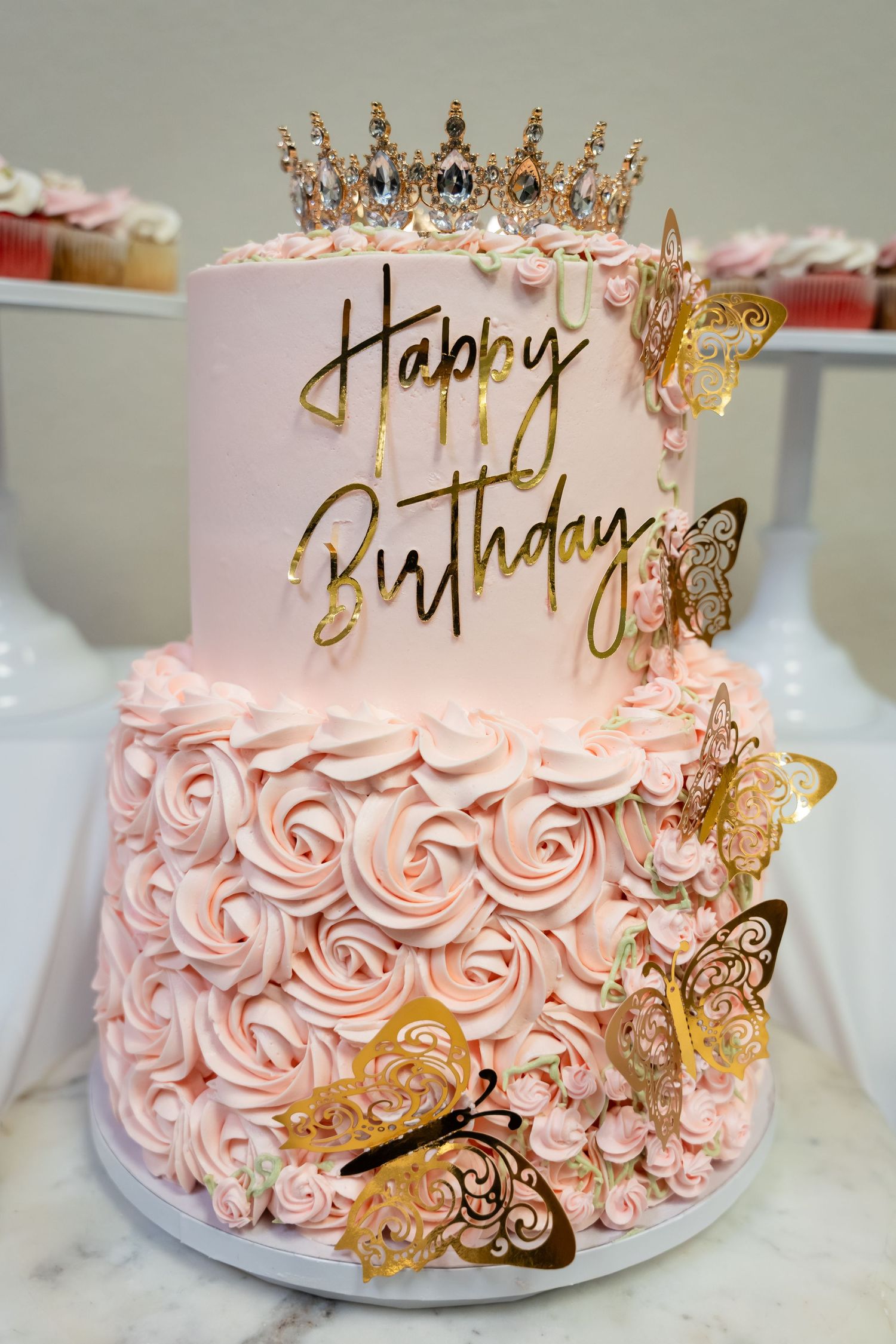 Two-tier pink rosette birthday cake with gold butterfly decorations, crown topper, and Happy Birthday script.