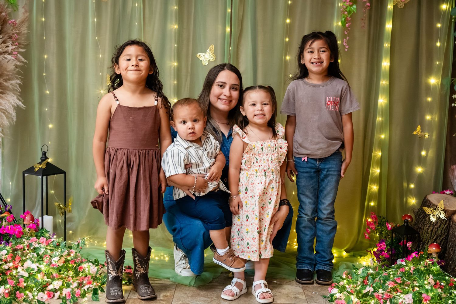 A woman kneels with four young children in front of a floral backdrop with fairy lights for a family portrait.
