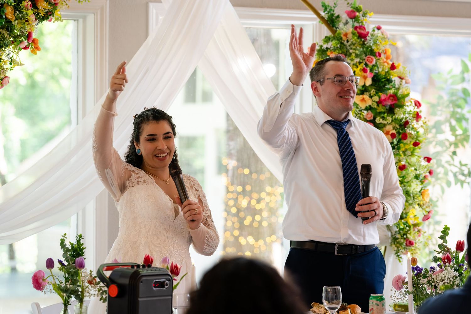 Bride and groom raising hands joyfully during wedding reception speech, standing under floral arch with white draping.