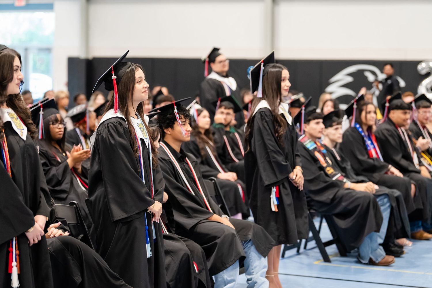 Students in black graduation gowns and caps stand and sit during a high school commencement ceremony indoors.