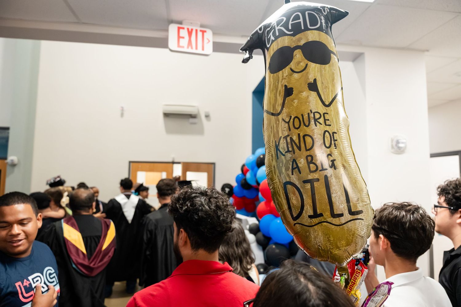 A festive graduation balloon shaped like a pickle reads 'You're Kind of a Big Dill' among students in gowns.