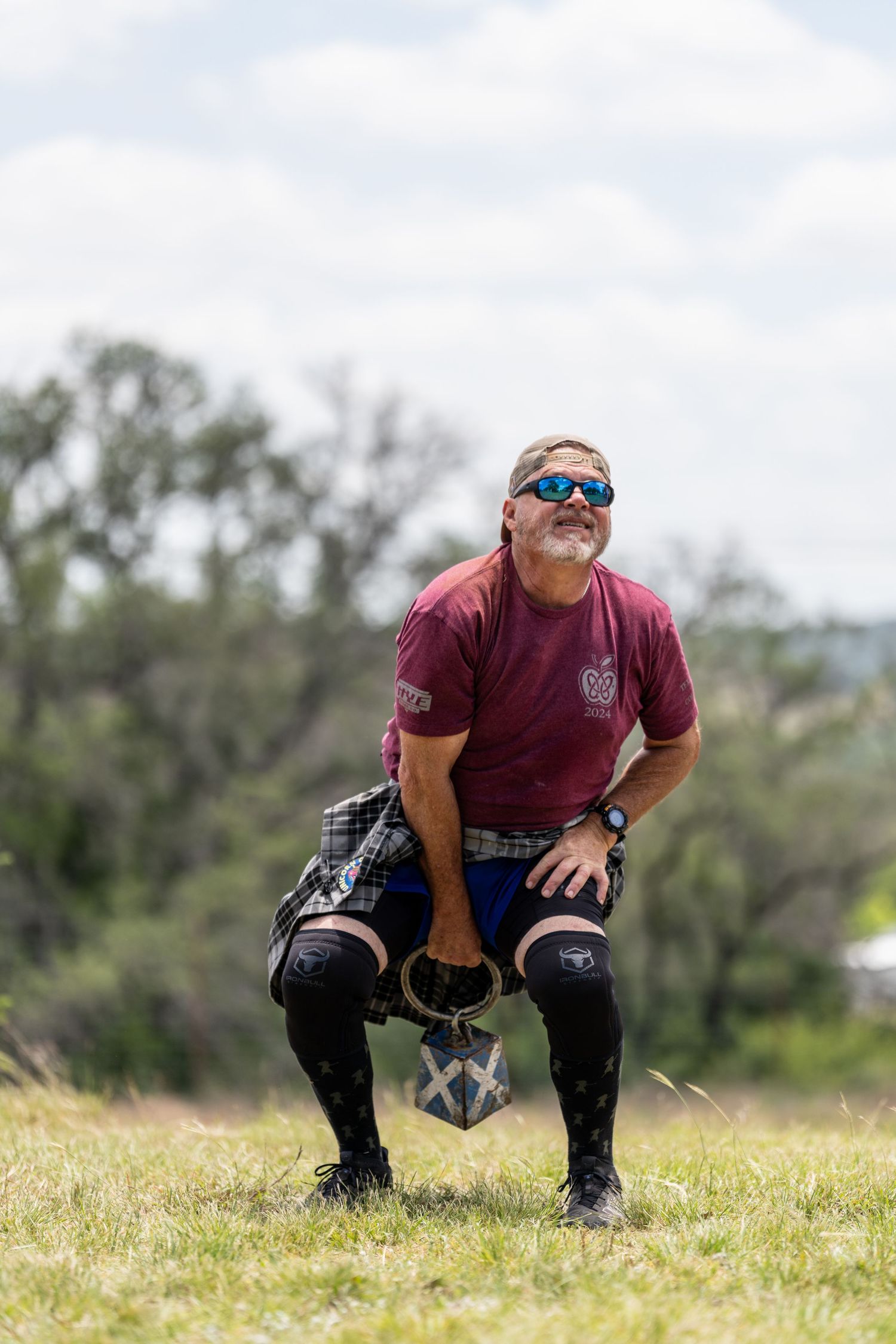 Man in kilt and maroon shirt crouches on grass field during Scottish Highland Games competition outdoors.