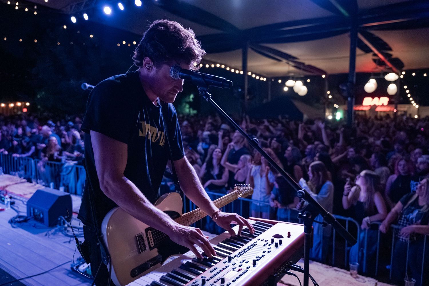 Musician plays keyboard and guitar on stage at an outdoor night concert with a large crowd in the background.