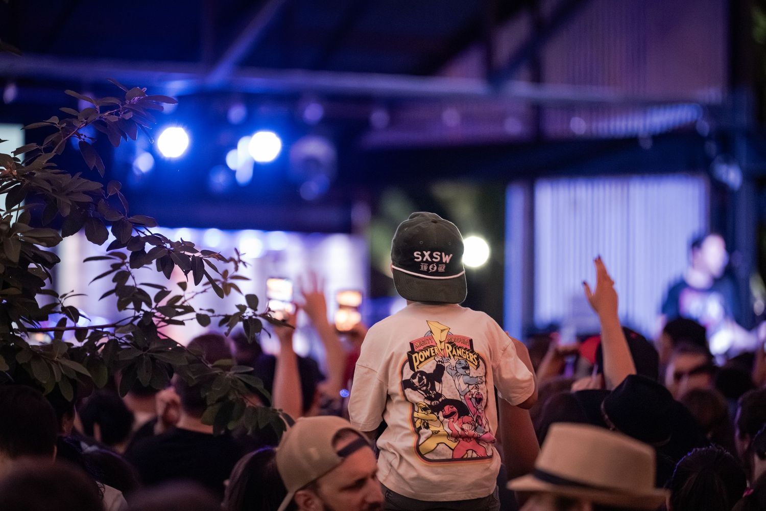 A child sits on shoulders above a crowd at an outdoor SXSW music festival concert with stage lights glowing.