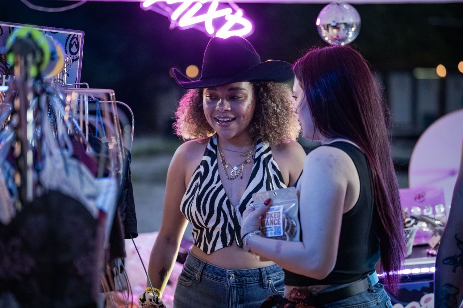 Two women chat at a neon-lit outdoor party, one wearing a cowboy hat and zebra-print top.