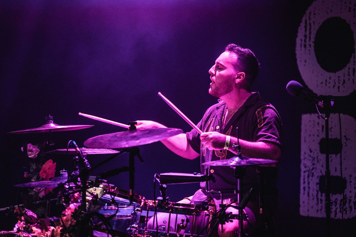 Drummer performs energetically on stage under dramatic purple lighting at a live concert performance.