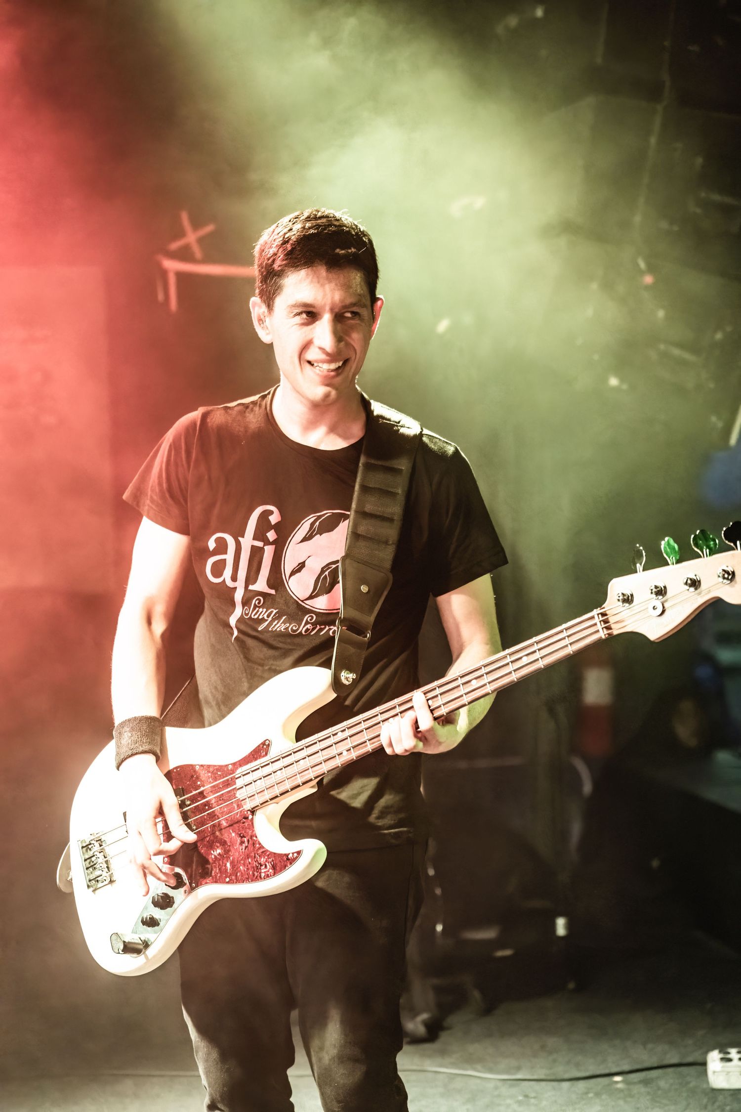 Musician playing white bass guitar performs on stage with red and green lighting effects.