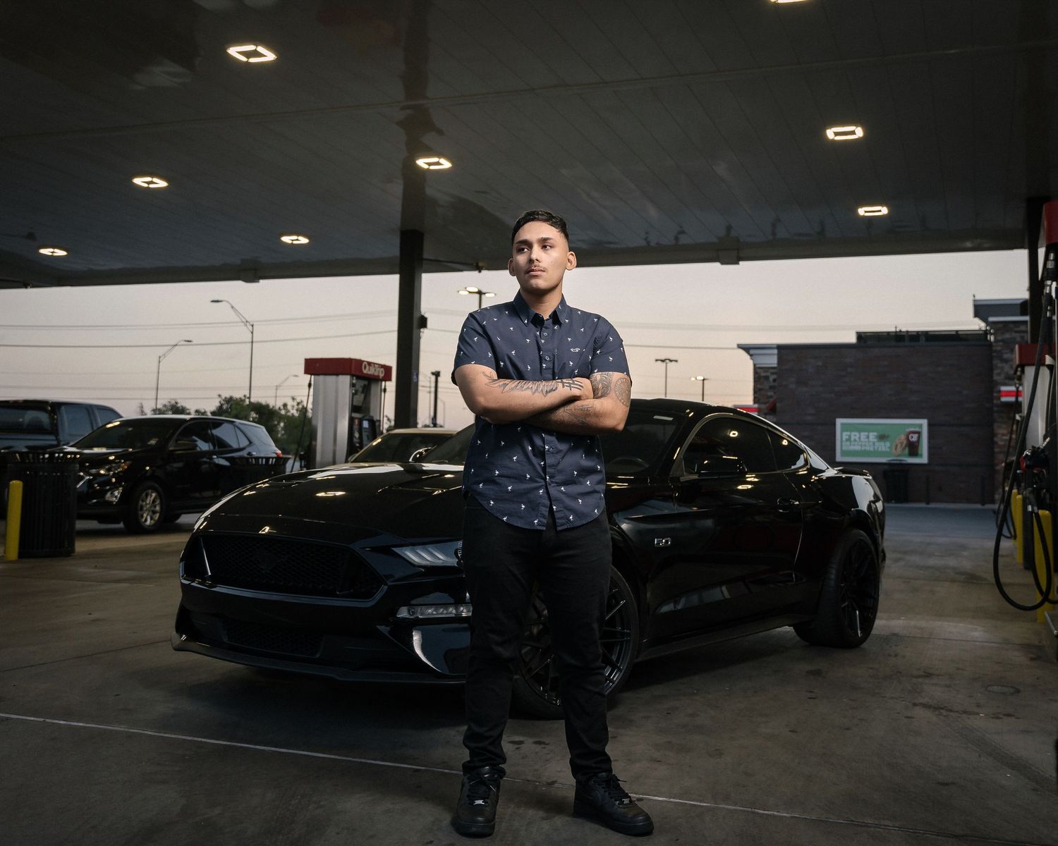 Person poses with arms crossed in front of black sports car in garage setting.