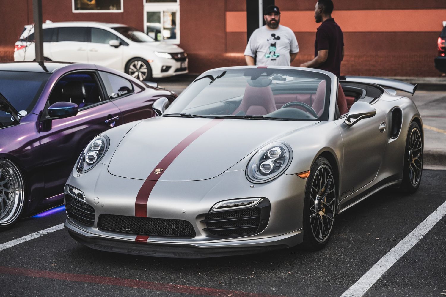 Silver Porsche 911 convertible with red racing stripe parked in lot amongst other luxury vehicles.