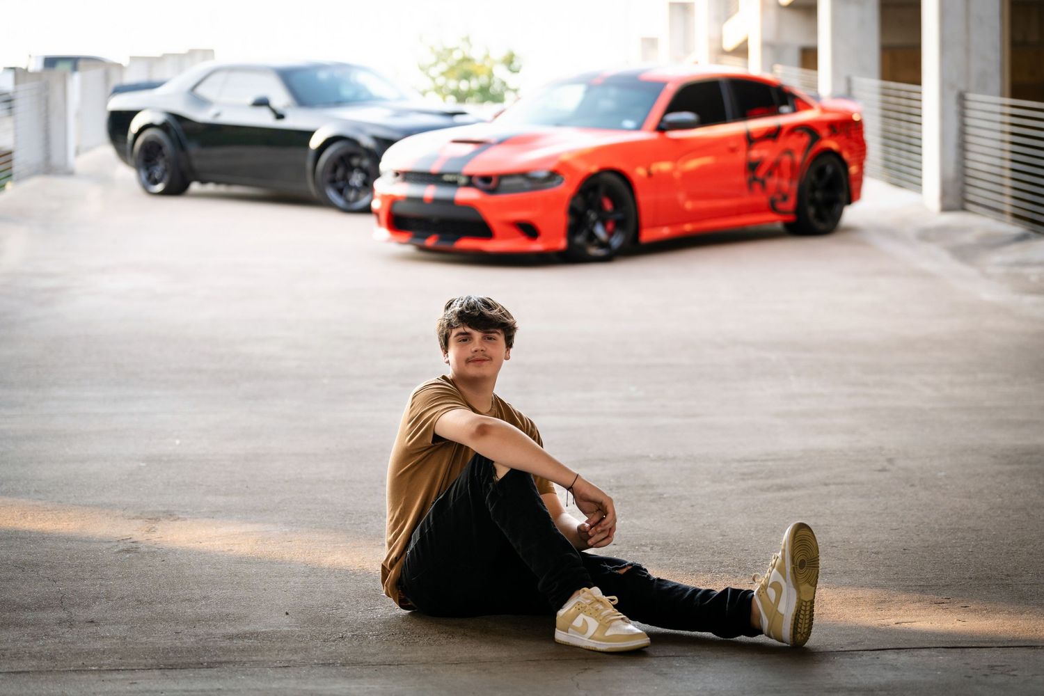 A brown car and orange sports car parked behind someone sitting casually on the ground.