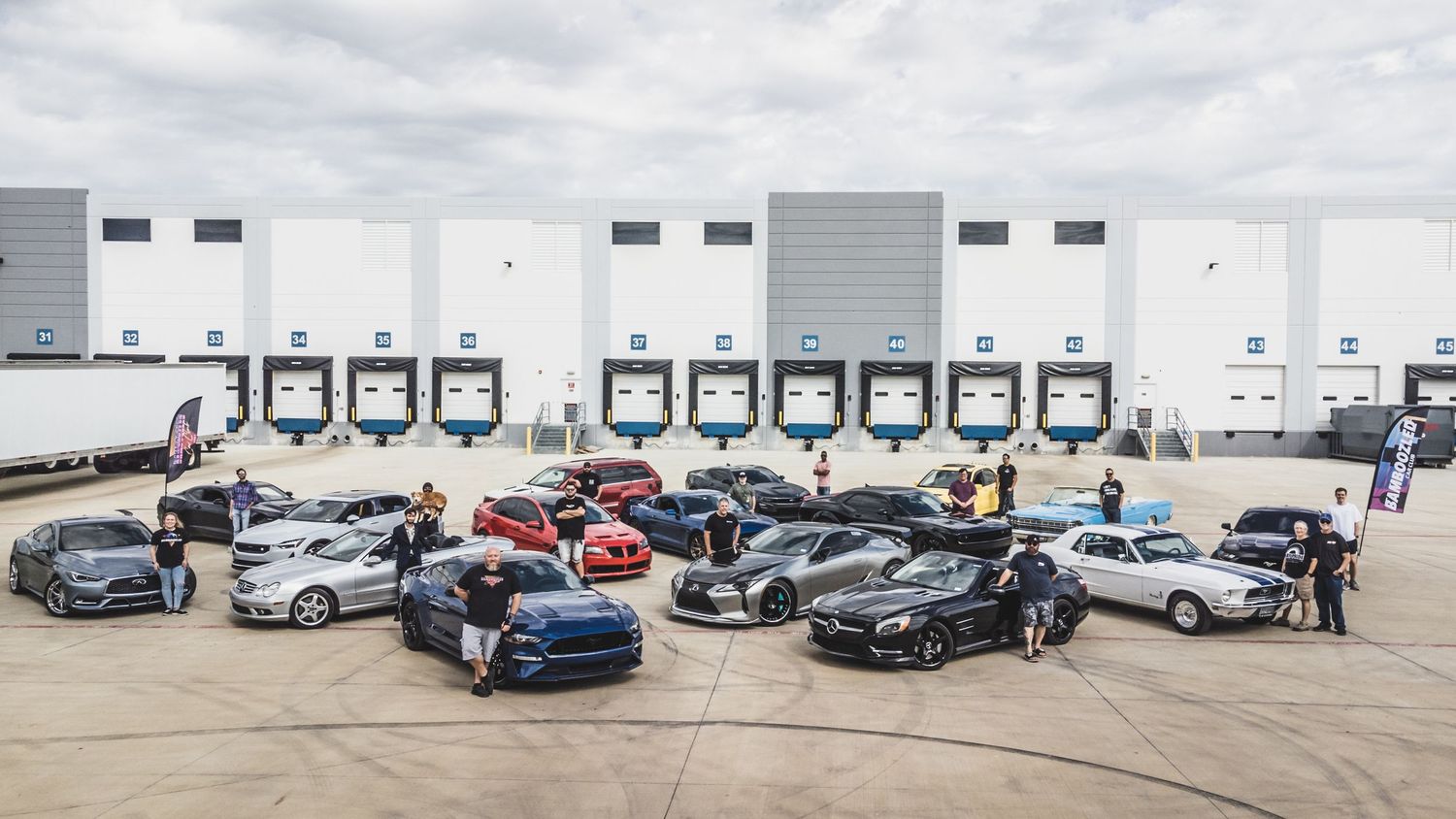 Car meet gathering showing diverse collection of vehicles parked at warehouse loading area.