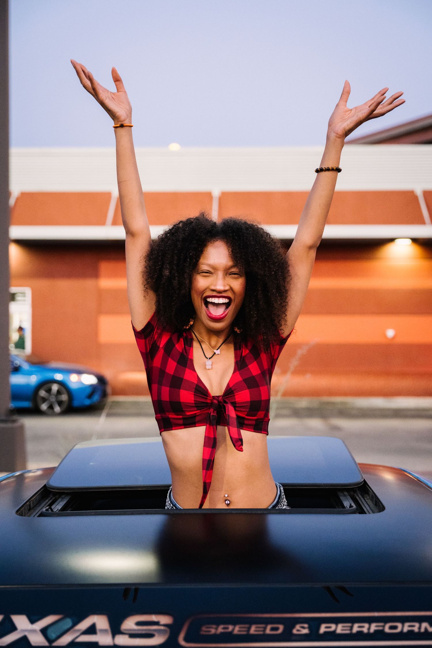 Person in red tied plaid top poses enthusiastically through car sunroof at outdoor event.