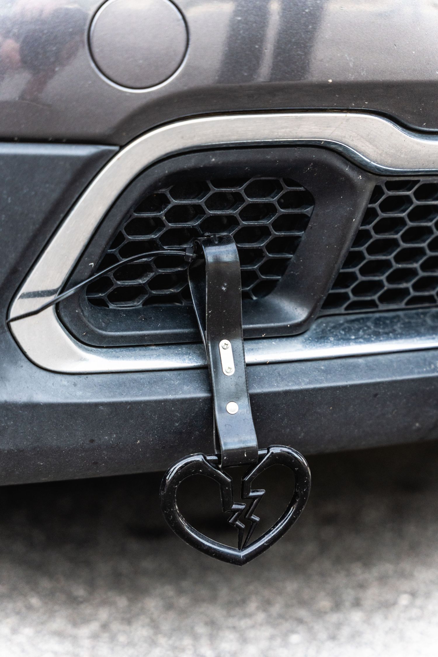 Close-up of a black heart-shaped ornament hanging from a vehicle's honeycomb grille.