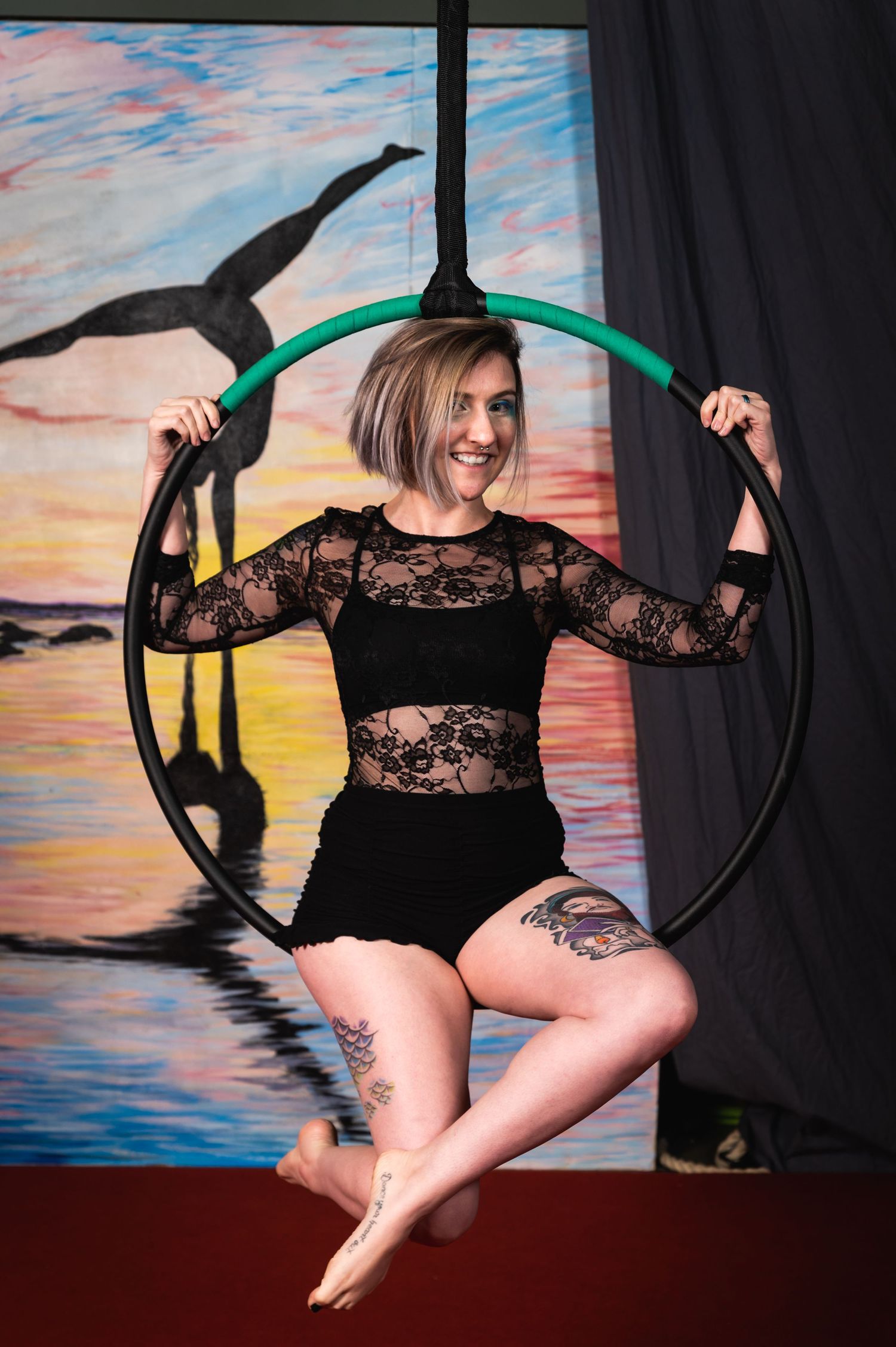 Performer poses gracefully in black lace outfit on aerial hoop against sunset backdrop.