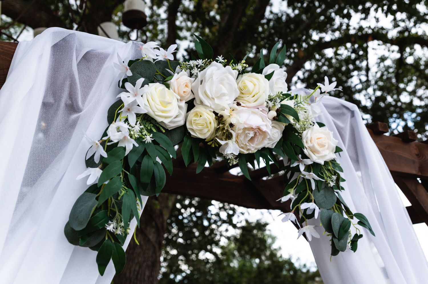 White roses and greenery adorning wedding arch with flowing sheer fabric.