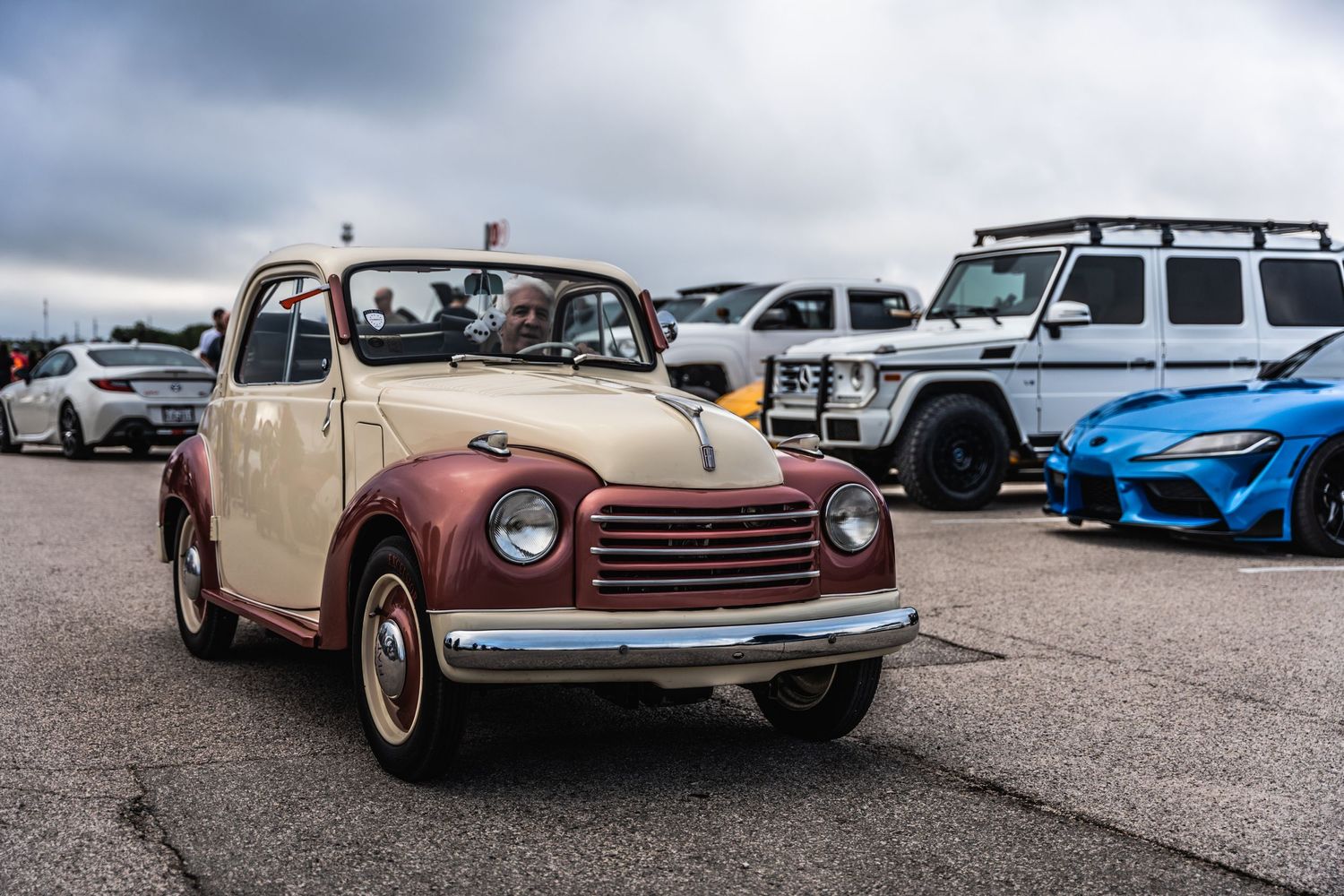 Vintage cream-colored Fiat 500 displayed among modern cars at automotive meetup.