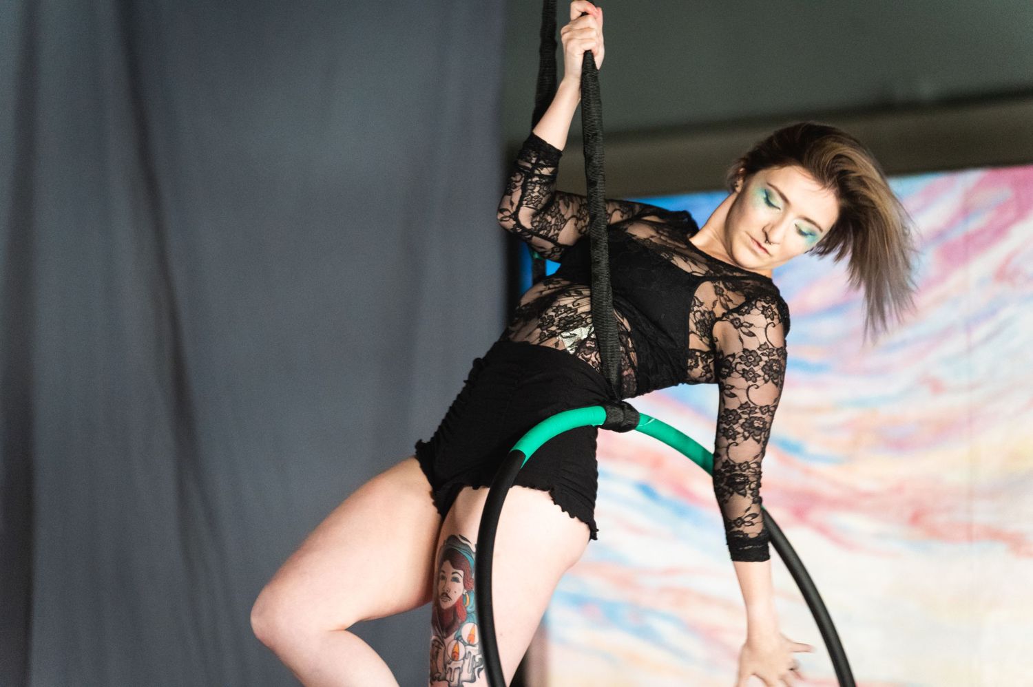 Aerial artist in black lace performs dynamic pose on suspended hoop against painted backdrop.