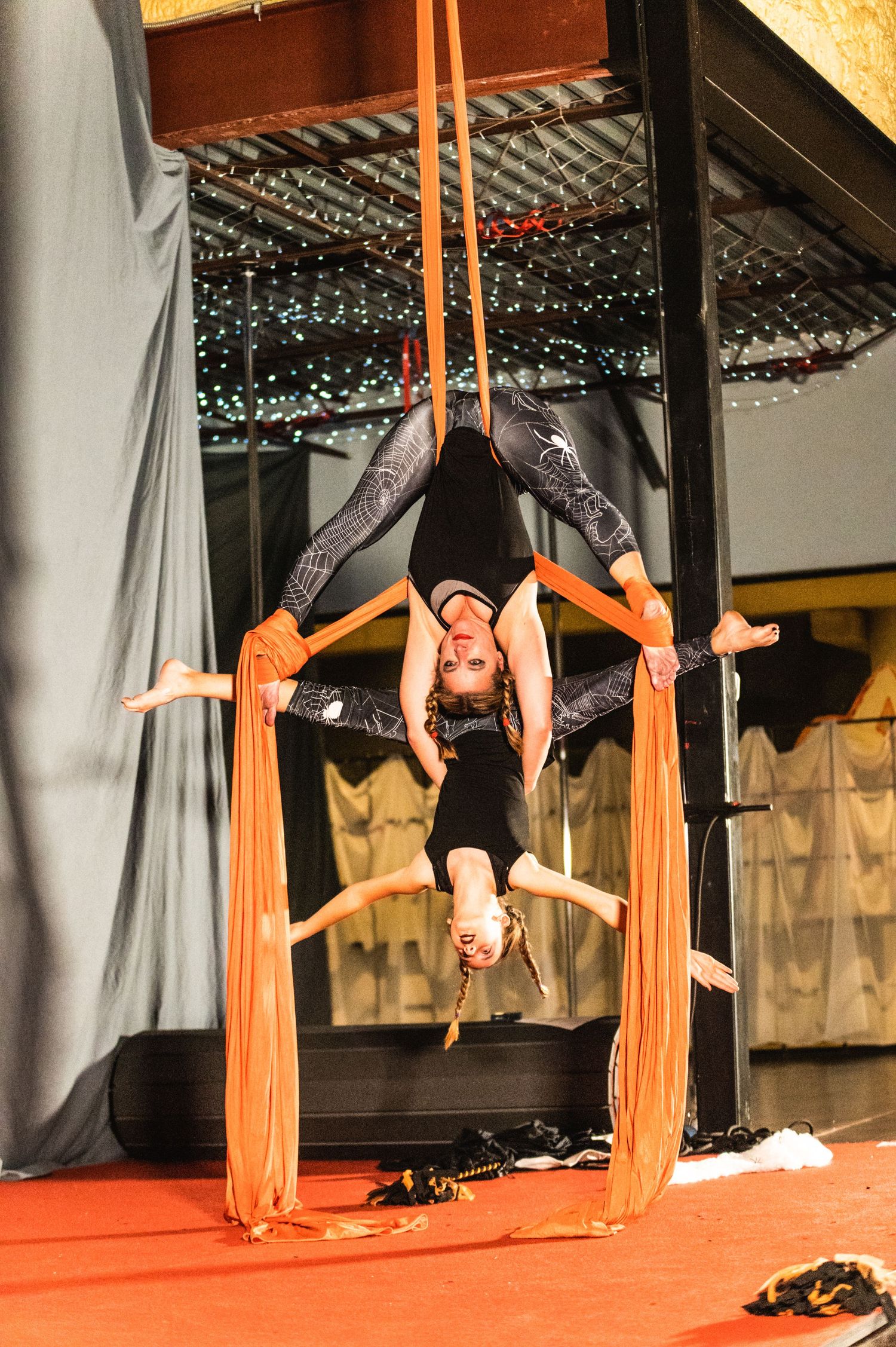 Aerial artist executes inverted pose between orange fabric streamers during performance.