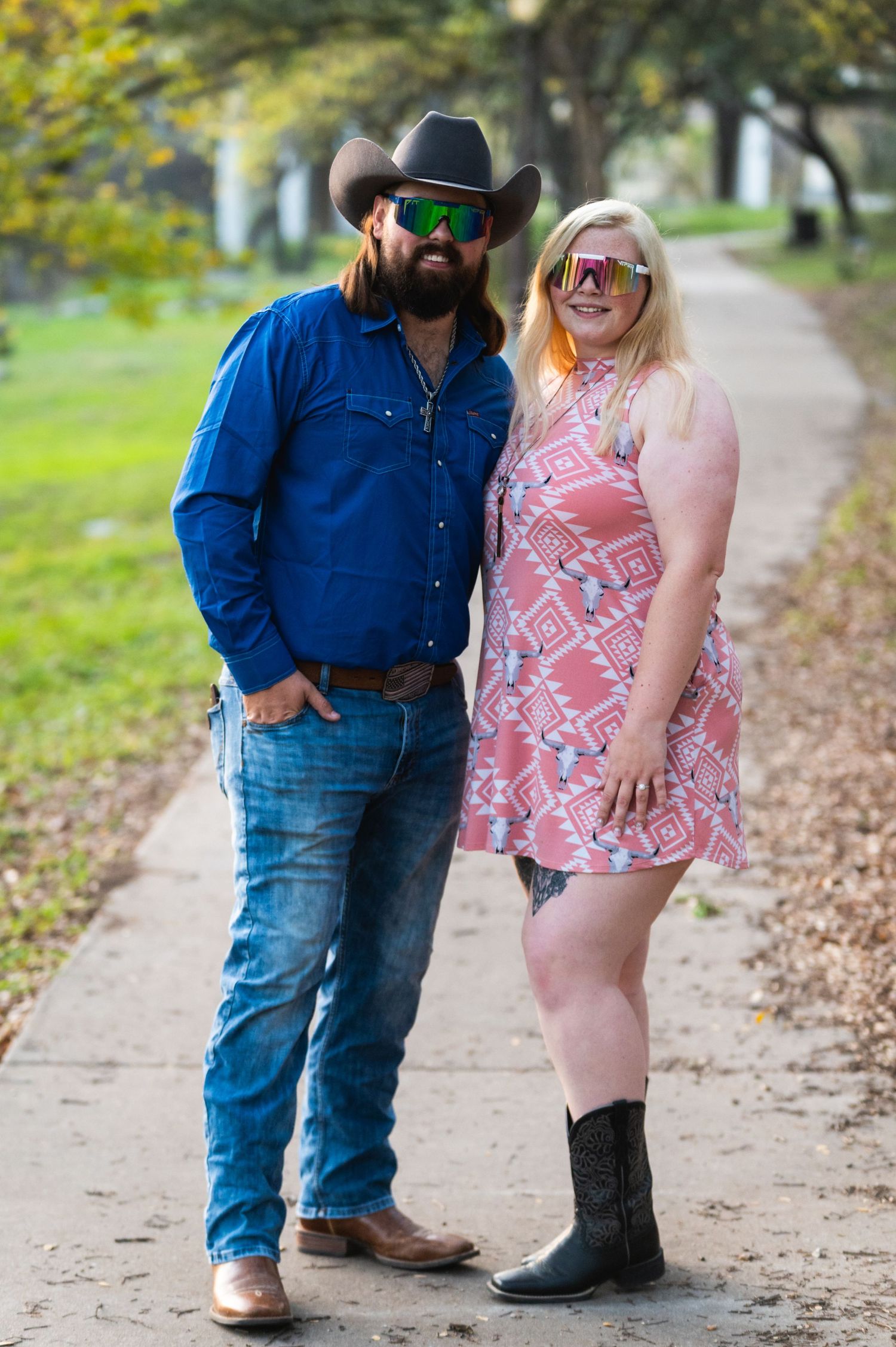 A couple in western wear smiles at the camera on an autumn path wearing coordinated blue and pink outfits.