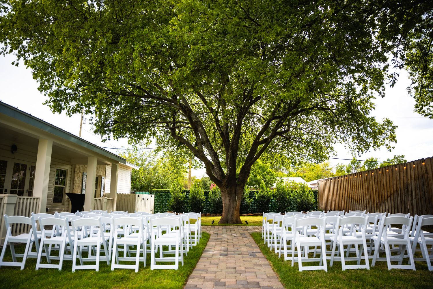 White chairs arranged for an outdoor wedding ceremony beneath a large shade tree in a garden setting.