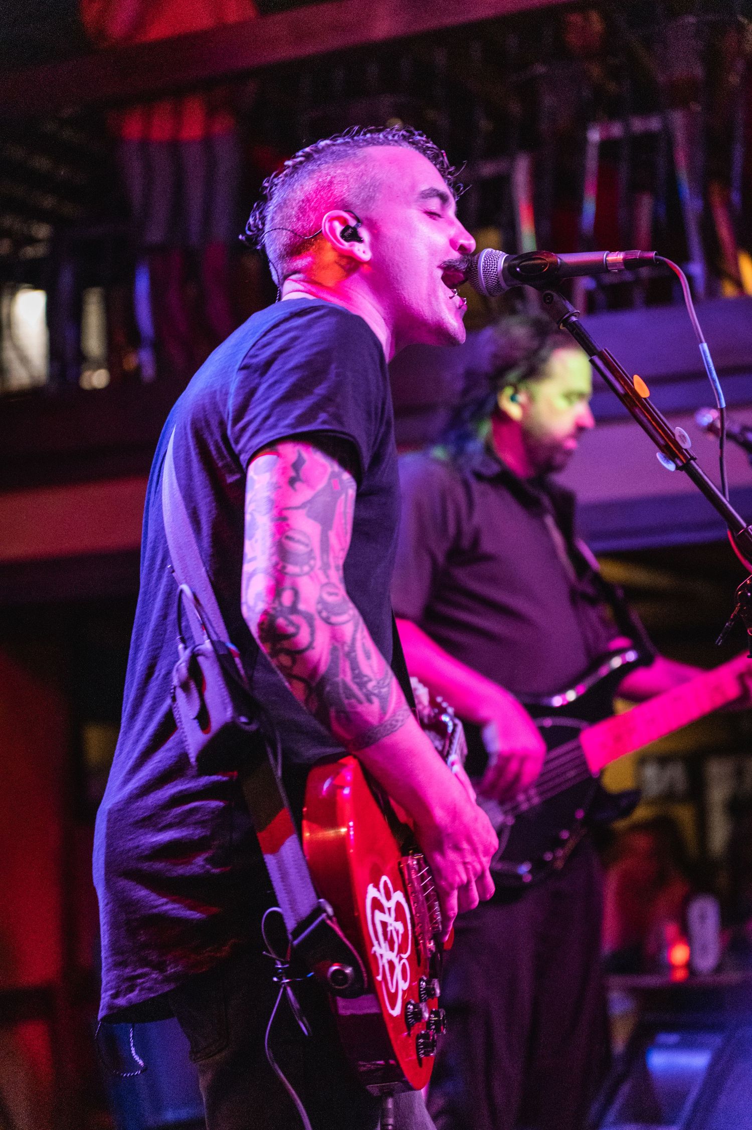 Tattooed musician plays red electric guitar and sings into microphone under pink stage lighting.