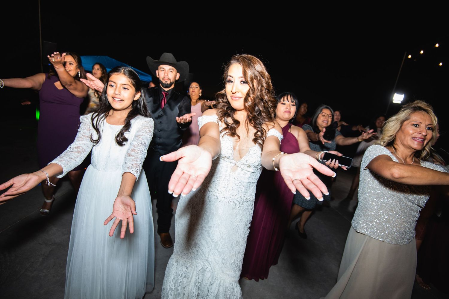 Wedding reception guests dancing in line with arms outstretched during nighttime celebration.