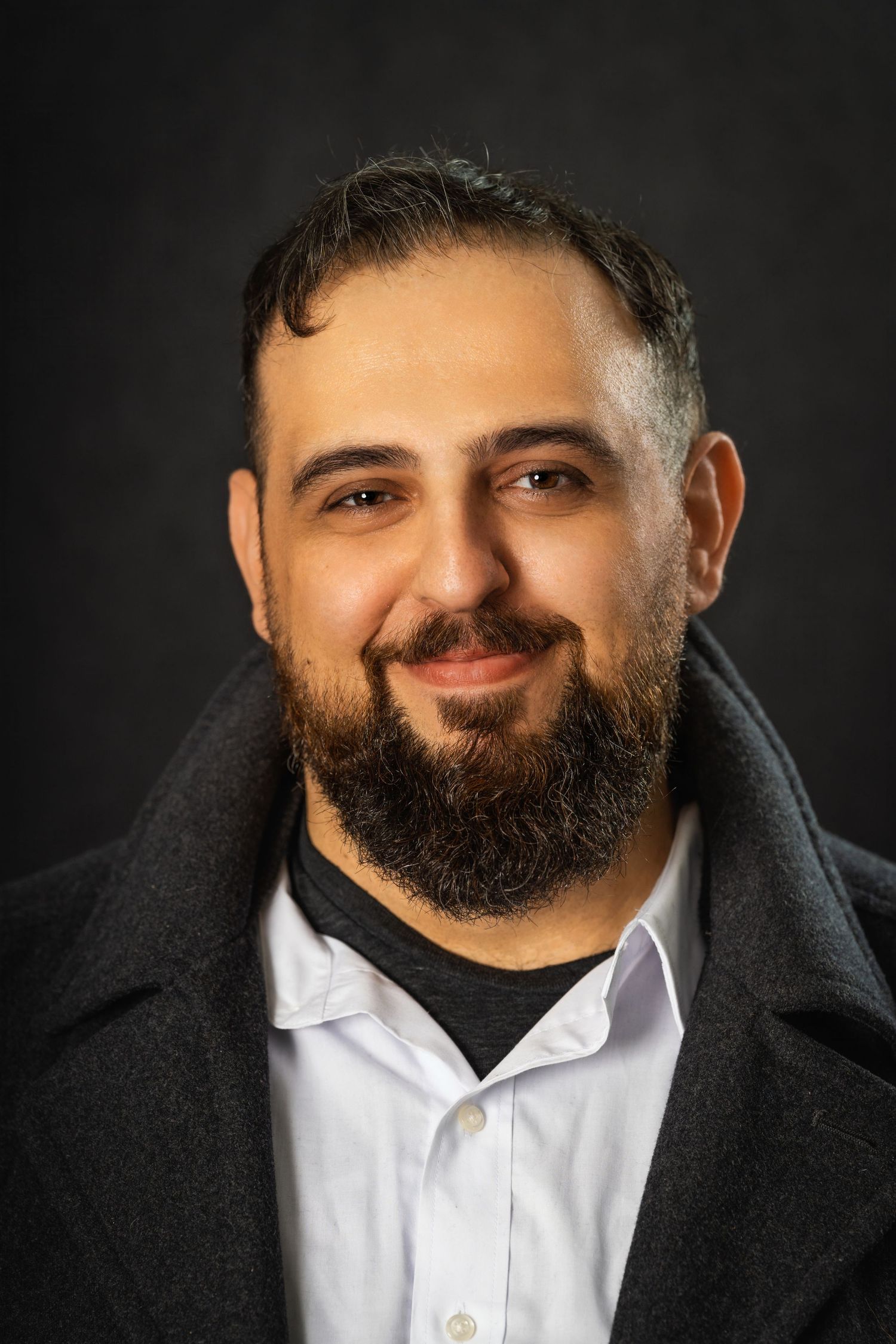 Professional headshot of bearded man in black coat against dark background.