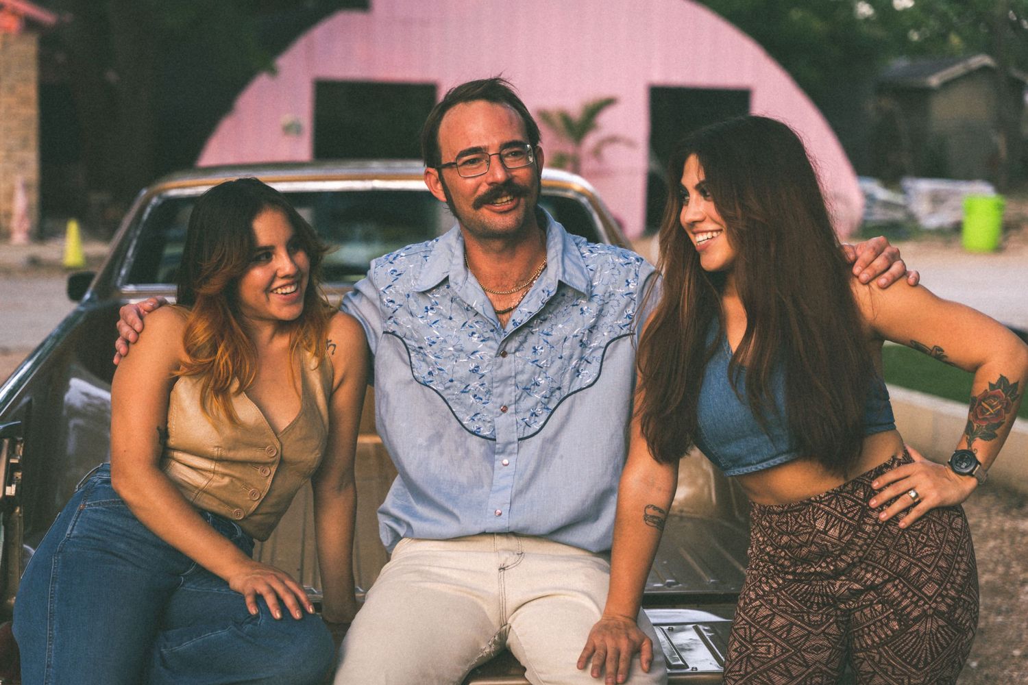 Three people laughing together while sitting on car hood during sunset.