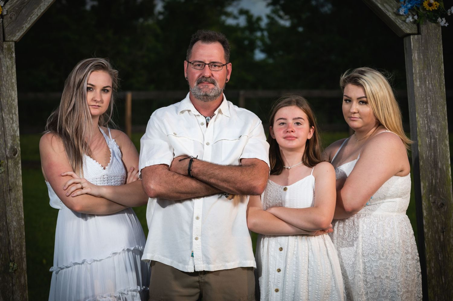 Family portrait of four people in white outfits posing under wooden arch outdoors.