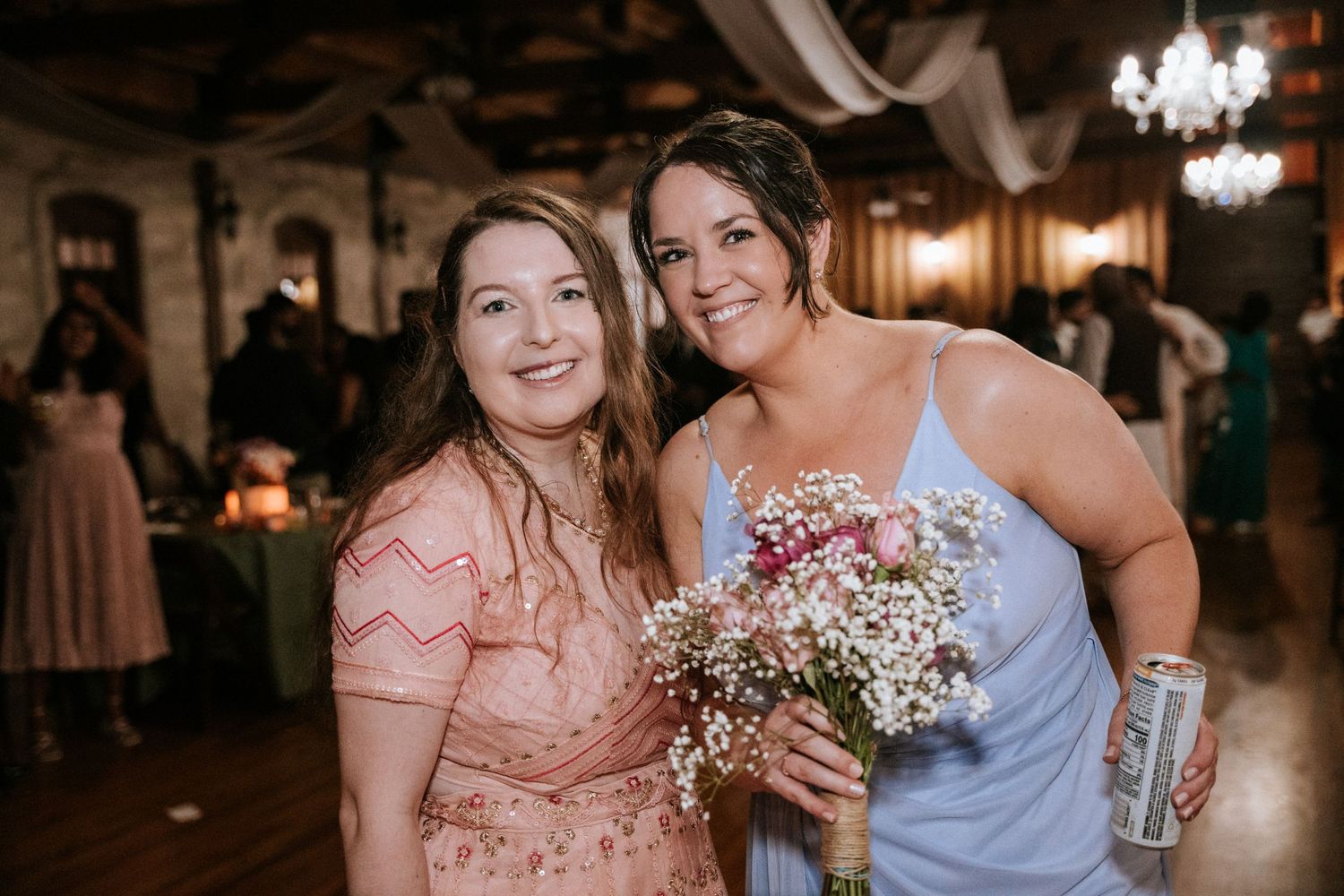 Two guests smile while holding a bouquet in a rustic venue with string lights.