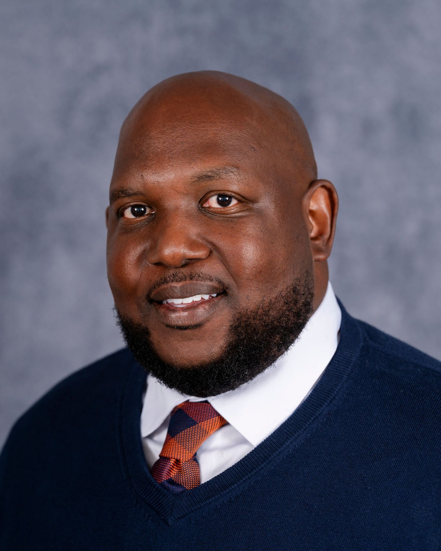 Professional headshot of person wearing navy sweater and red patterned tie against gray backdrop.