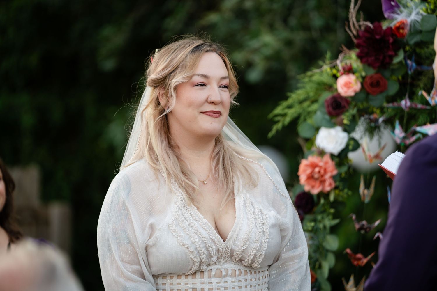 A bride in a white lace dress smiles during an outdoor ceremony with floral decorations.
