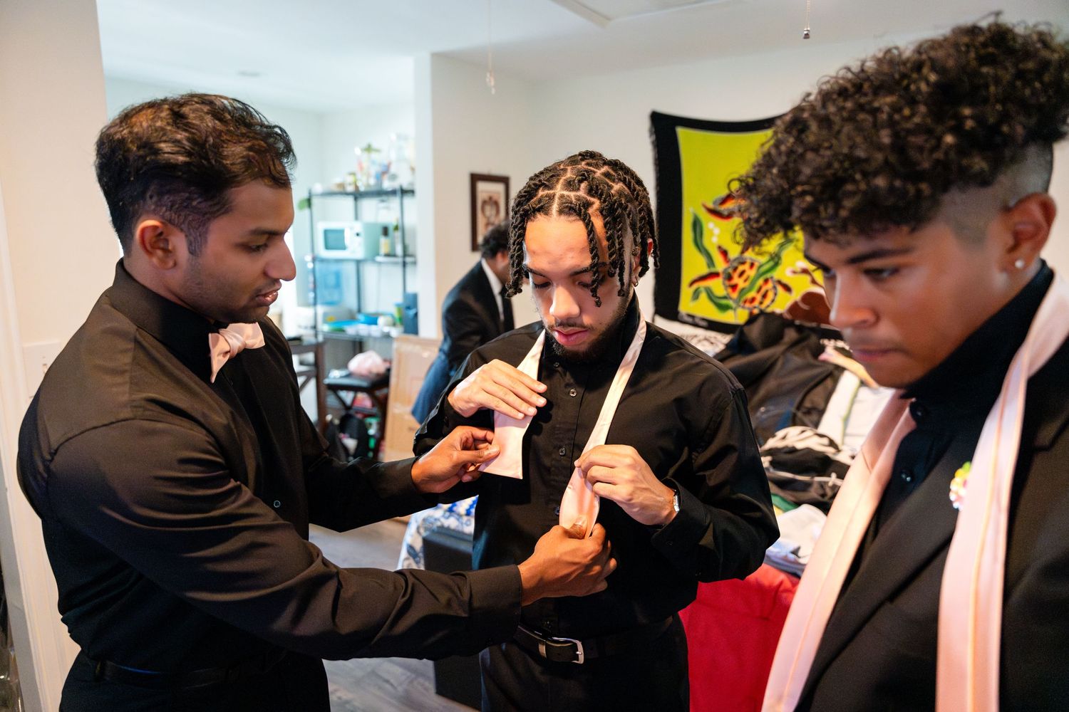 Groomsmen help adjust formal attire during wedding preparation in bright indoor setting.